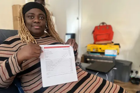 London heat network users say price cap lack has cost them dearly Dolly Thomas is holding a copy of her service charge bill, which has "estimated" written diagonally across it. Dolly is wearing a striped brown, black and red knitted dress and a black headband. She is seated in a chair inside her flat.