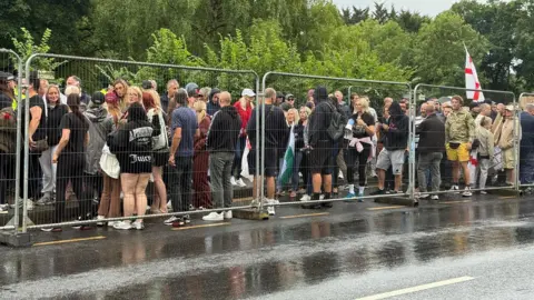 Epping council votes to urge government to shut asylum hotel Nadira Tudor/BBC A group of protesters, one holding an English flag, are stood on the either side of a metal fence on a rain soaked road.