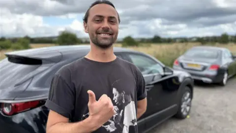Airport drivers try to beat drop-off charges by parking in laybys Rontom Tschopp smiling right at the camera. He is standing up but only the top half of his body is visible. He has his right thumb up to the camera. He has a short, dark beard and long, dark hair tied back. He is wearing a black T-shirt with a woman on it in black and white. He is standing in front of a black car and a silver car which are blurred in the background.
