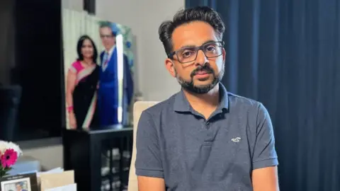 Victim's relative 'can't be at peace' until root cause known Miten Patel poses for a picture. He wears glasses and wears a blue polo shirt. A portrait of what appears to be his parents is seen in the background of the room.