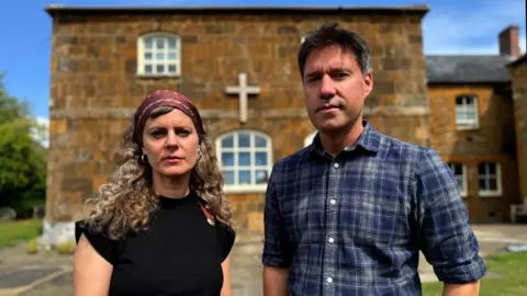 Investigating one of the UK's most abusive cults BBC Ellena Wood with long curly hair, a head scarf and a black top stands beside Jon Ironmonger, who is wearing a blue-checked shirt in front of a small orange-brick chapel building.