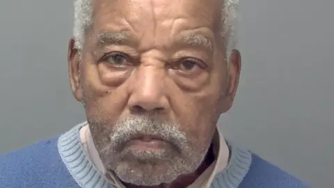 Cold case rapist and killer, 92, jailed for life Avon and Somerset Police Ryland Headley's custody picture. He has short white hair, and short facial hair, and wears a white collared shirt under a blue jumper. He is looking straight ahead with a serious expression.