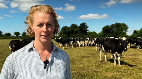 Dairy farmers face crisis as drought causes grass fails Dairy farmer stood in white checkered shirt. She has blonde hair, eye blues and is stood in front of cows standing in a field behind her.