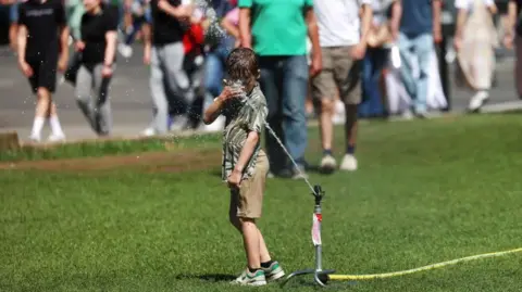 Hot weather continues as UK swelters past heatwave peak ANDY RAIN/EPA/Shutterstock A boy gets his head wet standing on grass in front of a water sprinkler at Parliament Square in London on 12 July.