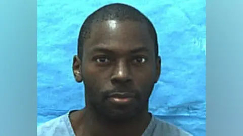 Florida Department of Law Enforcement Mugshot of Mark Gordon dated 15 January 2010. He is looking directly into the camera and is wearing a light blue top against a blue background. 