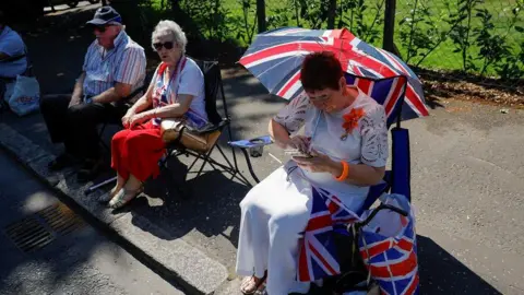 Thousands attending NI's Orange Order parades PA Media A spectator shading herself from the sun with a union jack umbrella. She is sitting down and wearing a while skirt and white top, she has short red-brown hair. To her right are a couple sitting on folding chairs. The woman is in a red dress and the man is in a striped red-white-and-blue shirt and dark blue cap.