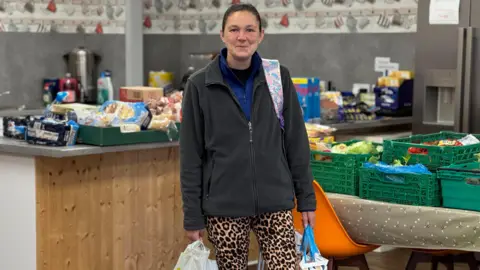 'Food demand in Cumbria is unprecedented' Leanne Hackett with dark hair tied back, wearing a black and blue fleece and leopard print leggings, holds two shopping bags as she stands in a kitchen full of boxes of food.