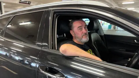 Airport drivers try to beat drop-off charges by parking in laybys Gavin Marshall sitting in his car looking directly at the camera. He has a greying beard and dark hair. His right arm is on the open window of the car, which is black. A parking garage is blurred in the background behind him.