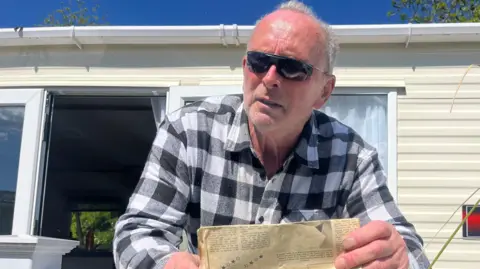 BBC Walter Renwick is standing looking over a white fence surrounding a static caravan, holding a faded yellow newspaper. He is a man in his 60s wearning a black and white checked shirt and sunglasses. 