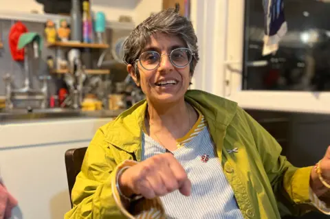 London heat network users say price cap lack has cost them dearly Sharon, who wears glasses and has greying hair, is sitting smiling in her kitchen with her first pointing towards the camera. Behind her is a washing machine and a sink. Above the sink is a blackboard in the shape of a cat.