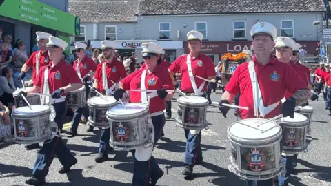 Thousands attending NI's Orange Order parades A parade of band members marching through Kesh wearing red short sleeved shirts, white hats and playing white drums. The sun is shining on the band members.