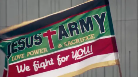 Investigating one of the UK's most abusive cults BBC/Docsville Studios The Jesus Army flag in green and red with a large red cross, reads 'Love, Power & Sacrifice; We right for YOU!'