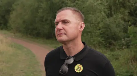 Battle of Orgreave national inquiry confirmed by Yvette Cooper A man standing on a rural footpath, with lots of trees behind him. He is wearing a black polo shirt, which bears a yellow badge reading 'coal not dole'. His sunglasses are tucked into the top of his shirt.