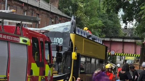 Fifteen hurt as double-decker bus hits bridge X A yellow double-decker bus has a smashed top windscreen and no roof, leaving the top floor seats exposed. A fire engine is parked in front of it and a man in a fireman's helmet can be seen stand on the street next to it speaking to people.