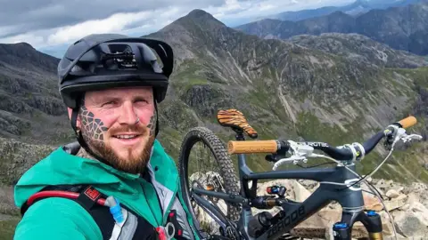 Mountain biker completes epic bid to ride down all 282 Munros Grizzly Munro Diaries A man who a tattooed face and a ginger beard wearing a mountain biking helmet. He is smiling, while standing next to a bike. Behind him is the view of a mountain, which shows he is high up.