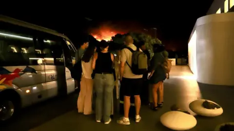 mass evacuations as blaze rages near Ierapetra, Crete EPA Tourists stand around next to a minibus outside a hotel after evacuating during a wildfire in Ierapetra, on Crete island
