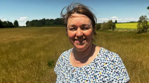 Dairy farmers face crisis as drought causes grass fails Prof Cannon stood in a spotty top in front of a field full of grass. The grass is yellowy brown.