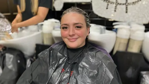 Package holidays to Spain, Cyprus and Turkey soar in price Abi Smitton / BBC News A woman at a hairdressers, with wet hair covered in conditioner. She's sitting in front of a sink and smiling at the camera. She's wearing a black cape and a bin bag over the top