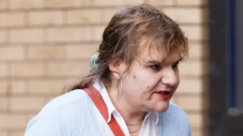 Ex-Tory MP given community order and fine for harassing ex-wife Athena Picture Agency A picture of a transgender woman with brown hair tied back, wearing dark eyeliner, lipstick, large hoop earrings, a white blouse and a blue cardigan, with a red crossbody handbag.