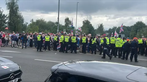 Hundreds of protesters gather at asylum hotel in Norwich area Richard Knights/BBC People waving flags and a line of police officers standing in front of them.
