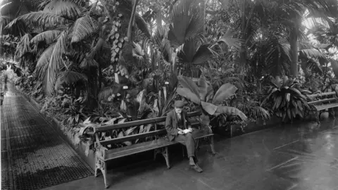 Palm House set for major renovation RBG Kew A black and white image taken inside the Palm House, a glass and iron building in Kew Gardens. Tropical plants fill the glass house and a man in a flat cap sits on a bench reading a book.