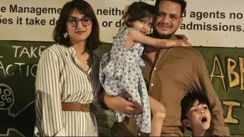 Families of Air India crash victims seek answers after report Javid Ali, hair short and wearing a brown button-down shirt, stands holding his daughter Amani, wearing a dress, beside his wife Maryam Ali, wearing a belted dress, glasses, and her dark hair short. Their son Zayn stands in front of them, Javid's arm around him.