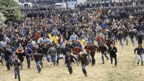 Battle of Orgreave national inquiry confirmed by Yvette Cooper Getty Images Dozens, possibly hundreds, of men run across a large patch of grass. Police officers, some of them on horses, can be seen in the background.