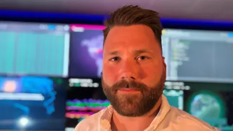 Small businesses tell of crippling costs of telecoms services BBC/Caroline Bilton Headshot of a man with short brown hair and a brown beard and moustache wearing a white shirt. He is stood in front lots of monitors.