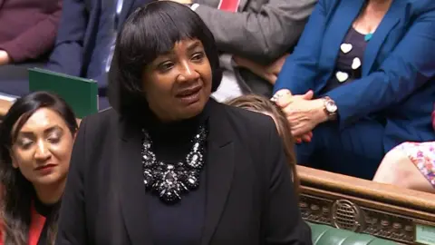 UK Parliament Diane Abbott stands in the House of Commons wearing a black outfit and a large statement necklace. She is surrounded by seated MPs dressed in various colors, including blue, grey, and red. The chamber’s green benches and ornate wooden paneling are visible in the background.