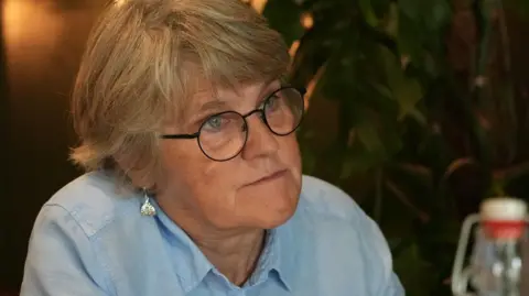 Tess McCay has short blonde hair.  She is wearing a light blue shirt and round glasses. 