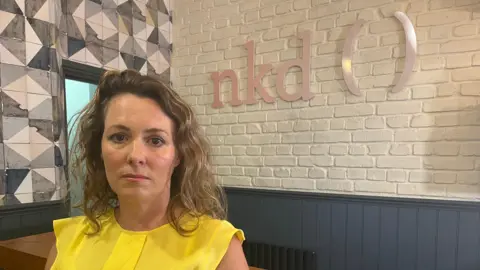 Salon owner 'ready' for key L'Oréal trademark dispute hearing BBC Woman wearing a bright yellow top standing in front of a sign which read nkd