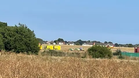 Southend Airport plane crash: Flights cancelled after incident Nick Hull/BBC A field with a number of ambulances and police cars parked in it. A row of houses is in the distance.