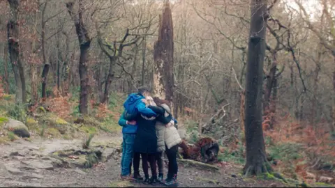 Investigating one of the UK's most abusive cults Docsville Studios A group of people in rain jackets give each other a group hug in a wooded area.