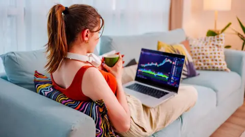 Getty Images Young woman sits on the couch with her feet up, coffee cup in hand, checking performance of shares on a laptop.