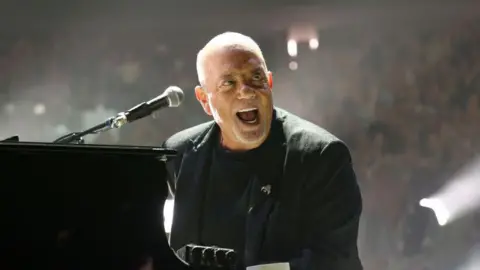 Billy Joel feels 'good' after brain disorder diagnosis Getty Images A bald man in a black t-shirt and black dinner jacket is performing on a piano, with a microphone pointed towards him. His mouth is open and smiling, with a crowd behind him, lit by spotlights.