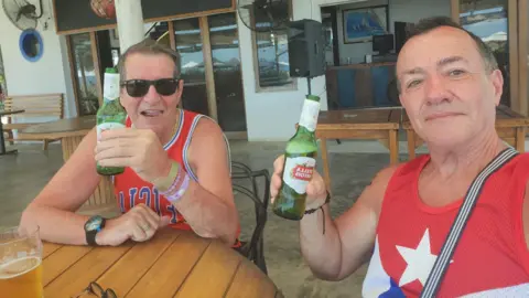 Extreme sex and bodies in suitcases Albert Alfonso/Flickr Mr Longworth, left, and Mr Alfonso, right, smile at the camera. They are both holding green bottles of Stella and wearing red vest tops with blue and white graphics on them. They are sitting at what appears to be an outdoor bar or restaurant, overlooking a view.