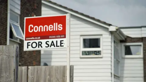 Dubious sales tactics at two leading estate agencies uncovered by BBC investigation Connells for sale sign - written in white letters on a red background - attached to a wooden fence. A house with white wooden cladding can be seen in the background.