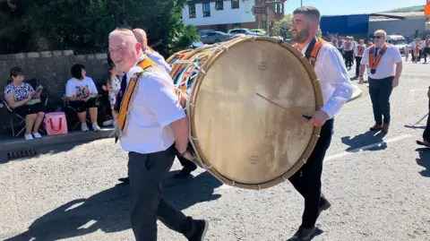 Thousands attending NI's Orange Order parades A parade with men carrying a lambeg drum on the street in the sun. Members are wearing white shirts and orange sashes.