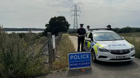 Man found guilty of murder of Anita Rose in Brantham in 2024 George King/BBC A police car is parked on a metalled track with a sign in front of it saying "POLICE" and "ROAD CLOSED". A police officer is next to the car, with two other people standing behind it. A pylon is in the field further along the track. The railway line and River Stour estuary are on the left of the picture.
