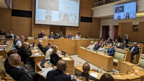 Epping council votes to urge government to shut asylum hotel Pete Walker/BBC The inside of a packed council meeting shows councillors sat at desks in a hall while other attend via large screens hanging overhead.