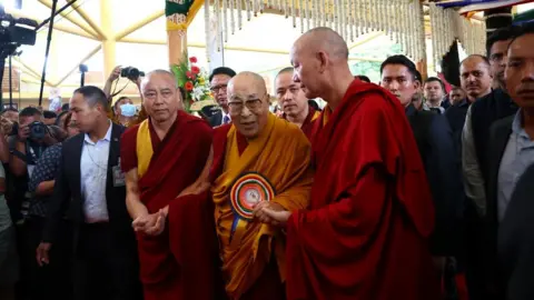 Thousands turn out to celebrate Dalai Lama's 90th birthday Reuters The bespectacled Dalai Lama cloaked in a red robe with a flowing orange wrap, arrives for his birthday celebrations flanked by two Buddhist's in red robes holding his hand. They are surrounded by suited men and a number of photographers.