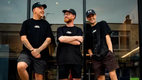 Porthcawl Coney Beach fair to close in October after 100 years Simon Hogarth Three Bone Cartel staff members outside the shop in black t-shirts with white letter branding and in black shorts and black caps. Simon Hogarth is in the middle and has a red beard