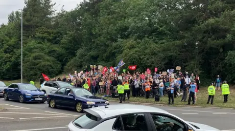 Hundreds of protesters gather at asylum hotel in Norwich area Alex Dunlop/BBC A large crowd of people standing by a junction. There is police around the group and protesters are holding flags.