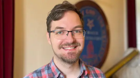230 years of unbroken weather recordings A close-up of Dr Rok Nežič smiling at the camera in Armagh Observatory. He has short, brown hair and a beard. He is wearing glasses and a red and blue checked shirt.