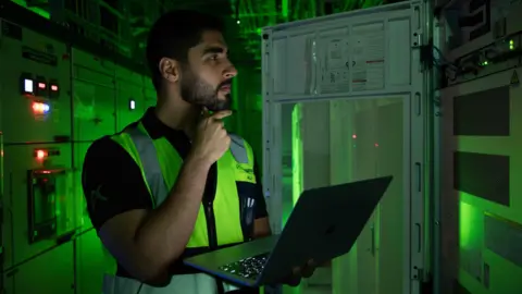 The Gulf bets big on AI as it seeks the 'new oil' Khazna A dark-haired man with a beard stands in front of a large computer in a data centre in the UAE. He holds an open laptop in his left hand and touches his beard with his right. The background is lit by a green glow from the surrounding equipment.
