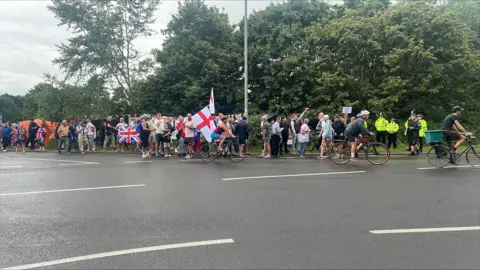 Hundreds of protesters gather at asylum hotel in Norwich area Richard Knights/BBC A large group of people holding the union jack and the St George 's flag. There are police officers at the scene.