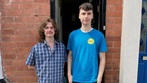No end in sight for Birmingham bin strike after six months Two men stood infront of a brick house, with a black doorway. The man on the left has long wavy brown hair, a moustache and is wearing a blue checked shirt. The man on the right has short brown hair and is wearing a blue T-shirt. Both are smiling at the camera.