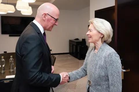 Trump back on the golf course for day two of Scottish visit European Commission's Audiovisual Service John Swinney, who has a bald head and is wearing a black suit jacket, shakes the hand of Ursula von der Leyen, who has short blonde hair and has grey suit jacket with a gold fastening.
