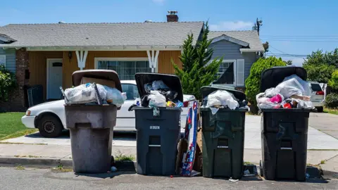 Getty Images Four overflowing bins stand on the curb in front of a small brown bungalow on a tree-lined street