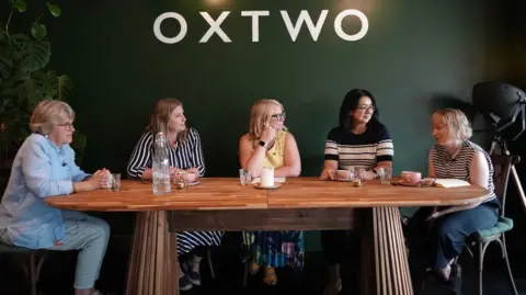 BBC Five women are sat around a dining table.  There pink coffee cups, a candle, water tumblers and a bottle of water on the table.